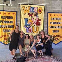 Six women pose in front of banners at Oxford Pennant Souvenir Shop, with one standing and five seated on a bench, smiling.