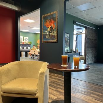Darkroom Brewing - a glass of orange juice on a table - A yellow armchair and a small round table with two glasses of drinks are in a lounge or cafe setting, with artwork on the wall and a view into another room. - Breweries Near Me - Geneva, Ohio