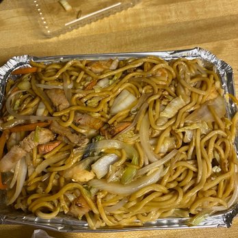 China One - House Lomein - A rectangular foil container contains house lo mein noodles mixed with vegetables such as onions, carrots, and cabbage, along with slices of meat. The container is on a wooden table, with some paper and plastic packaging nearby, indicating a takeout meal from a Chinese restaurant. - Chinese Near Me - Ripon, Wisconsin