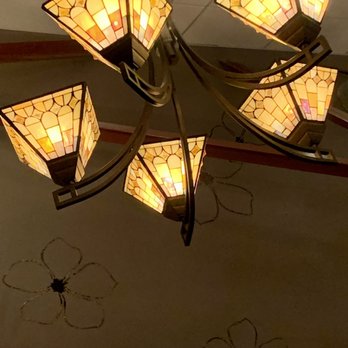 Adelle's - Decor - A chandelier featuring five stained glass lampshades in warm, amber tones hangs from a dark metal fixture. The lampshades have a geometric pattern and emit a soft, warm light. The ceiling has decorative floral outlines drawn with dark lines, and a small recessed light is visible in the background. - American Near Me - Wheaton, Illinois