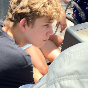 Village Pizza - Enjoying the patio - A young boy with curly blond hair is seated outdoors, leaning forward with his arms crossed on his knees. He appears to be focused on something in front of him. The background shows other people sitting on chairs, indicating a social or outdoor gathering. The image is slightly blurred, emphasizing the boy's concentration. - Pizza Near Me - Corrales, New Mexico