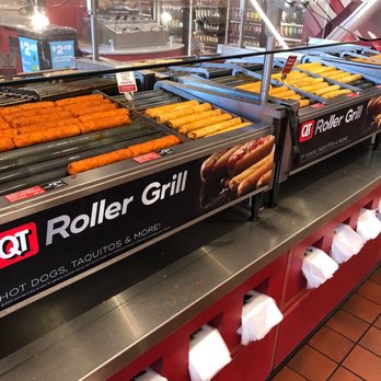 QuickTrip - Plenty of rollers. - The image shows a roller grill at a convenience store or fast-food outlet, with various hot dogs, taquitos, and similar snack items arranged on rotating metal rollers. The grill is covered by a glass sneeze guard, and there are price signs indicating $2.49 for some items. The background includes shelves stocked with beverages and other products, and the counter has paper napkins stored underneath. The setting appears to be a typical convenience store or quick-service food area. - Gas Stations Near Me - Mesa, Arizona