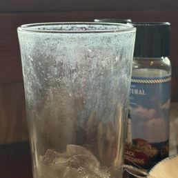 Dirty glasses! - A tall, transparent glass contains ice cubes and has visible condensation on its exterior, indicating cold temperature. Behind the glass, there is a bottle of mineral water with a label featuring a landscape scene, including clouds and a coastal view. The setting appears to be indoors near a window, with natural light illuminating the scene.