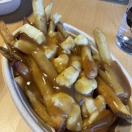 Classic - French fries topped with cheese curds and brown gravy are served in a disposable paper bowl on a wooden table. The dish appears to be a poutine-style snack, with crispy fries and melted cheese curds covered in gravy. In the background, there are other food items and a glass of water, indicating a casual dining setting.