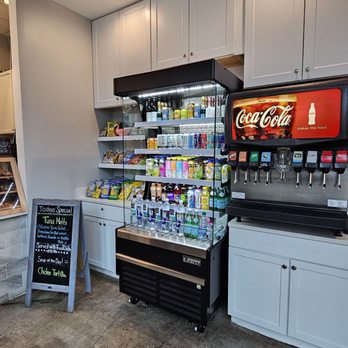 Uptown Cafe - A beverage refrigerator filled with bottled water and assorted drinks is situated next to a Coca-Cola soda fountain with multiple dispensing nozzles. The refrigerator is positioned against white cabinets with closed doors, and a small chalkboard sign with daily specials is nearby. The floor is concrete with visible stains, and ceiling lights illuminate the area. - Coffee & Tea Near Me - Chantilly , Virginia
