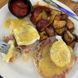 Eggs benny - The image shows a breakfast plate with two eggs Benedict topped with hollandaise sauce, served on bread with ham. Accompanying the eggs are roasted potatoes with a slightly crispy exterior. A small black container of ketchup is also on the plate. The meal is placed on a wooden table, with a butter knife and a glass of a light-colored beverage nearby.