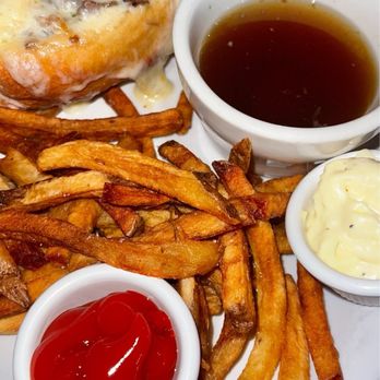 Johnny Seesaw's - Prime Rib French Dip - A French dip sandwich topped with melted cheese, served with French fries and small bowls of ketchup, mayonnaise, and au jus on a white plate. - American Near Me - Peru, Vermont