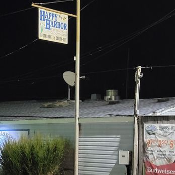 Happy Harbor - The image shows a sign for Happy Harbor restaurant and bar, mounted on a tall pole outside a building at night. The sign displays the business name, phone number, and indicates it offers restaurant and carry-out services. The building has a satellite dish on the roof and a poster advertising drinks and domestic wine. Vegetation and electrical wires are also visible in the scene. - Venues & Event Spaces Near Me - Deale, Maryland