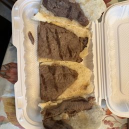 Steak sandwich - Four beef steak sandwiches with grilled beef and bread are arranged in a white takeout container on a floral tablecloth.