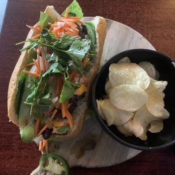 Wicked Pho - banco  Beef Sandwich - A beef banh mi sandwich topped with fresh vegetables and cilantro, served with a bowl of potato chips on a wooden board. - Vietnamese Near Me - El Paso, Texas