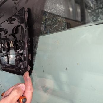 A Better Windshield - A person’s hand, holding a screwdriver or similar tool, points at the inside of a car windshield. The image shows the interior side of the windshield with some visible mounting or repair components. The background includes reflections and parts of the vehicle’s interior, indicating a repair or inspection process. - Windshield Installation & Repair Near Me - Monument, Colorado