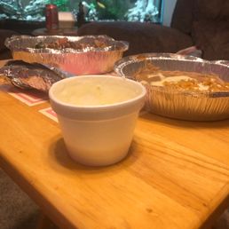 a table with pies and a bowl of soup - Several aluminum foil containers with food and a white cup are arranged on a wooden table. The containers appear to hold different dishes, and the cup contains a creamy or frothy substance. In the background, a fish tank with aquatic plants and fish is visible, along with a brown couch and some objects on a side table. The setting suggests a casual dining or gathering area.