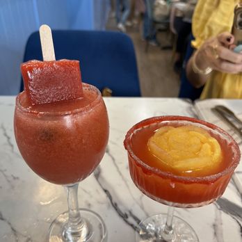Fiore - Drinks - The image shows two red-colored drinks on a white marble table in a restaurant. The drink on the left is in a tall glass with a popsicle on top, while the drink on the right is in a coupe glass with a yellow fruit garnish. The background features a busy restaurant with people dining and a woman in a yellow top holding a phone. - New American Near Me - Miami, Florida