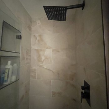 Established Homes - Stall shower - The image shows a shower area with beige marble-like tiles on the walls. A black rain shower head is mounted on the ceiling, and a black wall-mounted control handle is visible. There is a built-in wall niche with two shelves containing toiletries such as bottles and containers. The lighting is dim, and the overall design appears modern and minimalistic. - General Contractors Near Me - East Lyme, Connecticut