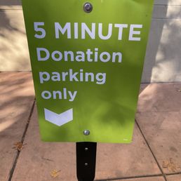 The image shows a green, rectangular sign mounted on a black post on a sidewalk. The sign has white text that reads, '5 MINUTE Donation parking only,' with a downward-pointing arrow below the text. The background includes a concrete wall and some fallen leaves on the sidewalk, indicating an outdoor setting.