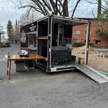 The Ranch Hand - Outside - A black food truck with a serving window and a wooden table outside in a parking lot, with trees and a brick building in the background. - Food Trucks Near Me - Chico, California