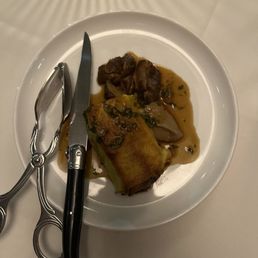 Short Rib Cannelloni - A white plate holds a rolled food item with sauce and small meat chunks, set on a white surface. Scissors and a knife are placed on the plate.