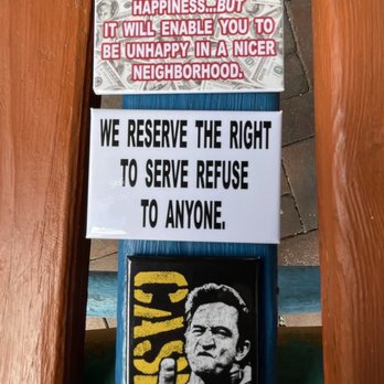 Guerrilla Graphix - Three signs are arranged vertically on a wooden bench with blue and orange painted slats. The top sign features a humorous message about happiness and neighborhood, with a background of dollar bills. The middle sign states a political message about the right to refuse service, in bold black text on a white background. The bottom sign is a black and yellow stencil-style image of a man making a rude gesture, with the word 'BASH' partially visible behind him. The setting appears to be outdoors, with a paved surface visible beside the bench. - Printing Services Near Me - Albuquerque, New Mexico