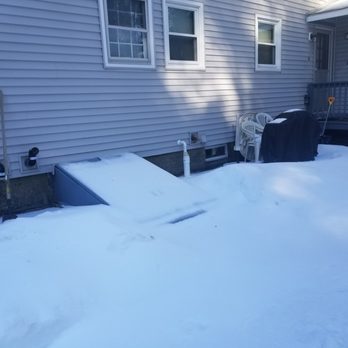 Ulster Property Services - The image shows a snow-covered backyard of a house with vinyl siding exterior. Snow covers a small ramp leading to a basement door, patio furniture including chairs, and a grill with a cover. A shovel is leaning against the house near the deck. The house has three windows and a vent pipe visible on the wall. Shadows from nearby objects are cast on the siding. - Landscaping Near Me - Bedford, New Hampshire