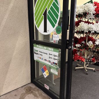 Dollar Tree - The image shows the glass entrance door of a Dollar Tree store, featuring the store's logo and various stickers, including Uber Eats and other notices. The store hours are posted, indicating opening times from 8 am to 10 pm Monday through Saturday and 8 am to 10 pm on Sunday. To the right of the door, there is a display of red and white artificial flowers on a stand. The door is set in a beige wall, with a concrete sidewalk at the base. - Discount Store Near Me - Placentia, California