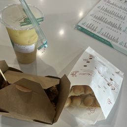 The image shows a white table with a cup of green tea yogurt drink with a straw, a paper bag containing cream-filled buns, and a printed menu. The drink is in a clear plastic cup with a sealed lid, and the buns are in a white paper bag with decorative text. The menu is partially visible, with multiple items listed, suggesting a cafe or casual dining setting.