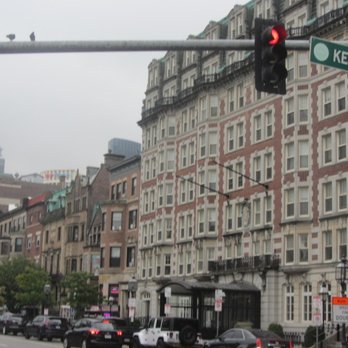 Kenmore Square - The image depicts a city street corner in Boston, Massachusetts, featuring multi-story buildings with architectural details, a traffic light showing red, and several cars parked along the street. A street sign indicates Kenmore Street, and the scene includes trees and urban infrastructure, capturing a typical cityscape environment. - Landmarks & Historical Buildings Near Me - Boston, Massachusetts