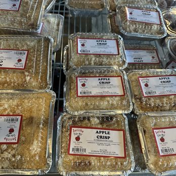 Jericho Cider Mill - Assortment of cakes and pies - The image shows several aluminum containers of apple crisp with labels, arranged on a metal shelf at Jericho Cider Mill. The labels indicate the product name, ingredients, and business details, including the address in Jericho, NY. The containers are stacked and positioned in a retail setting, with a rustic wooden background and some plastic cups visible on the side. - Fruits & Veggies Near Me - Jericho, New York