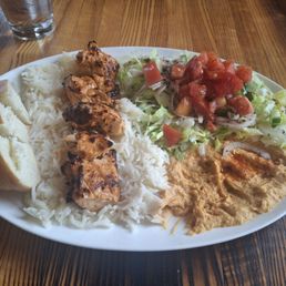 Chicken shish kabob platter - A plate of Turkish cuisine features grilled chicken pieces on a bed of white rice, accompanied by a fresh salad with chopped tomatoes, cucumbers, and onions, topped with tomato sauce. There is also hummus with a reddish-orange spice or sauce and a piece of bread on the side. The meal is served on a white oval plate placed on a wooden table, with a glass of water visible in the background.