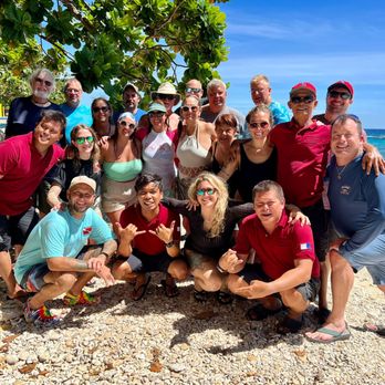 Force-E Scuba Centers - A group of approximately 20 people is gathered on a rocky beach, with some standing and others crouching in front. They are smiling and appear to be enjoying a sunny day. The background features the ocean with visible waves and a clear blue sky. Some individuals are wearing sunglasses, hats, and casual summer clothing, indicating a warm climate. The scene suggests a social gathering or vacation at a seaside location. - Scuba Diving Near Me - Boynton Beach, Florida