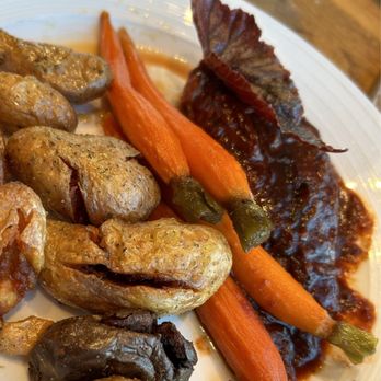 Devil's Kitchen - Chicken - A white plate contains roasted carrots, chicken pieces, and a piece of meat topped with a dark sauce. The carrots are whole and cooked, with green stems attached. The meat appears to be a cut of beef or similar, covered in a dark sauce. The dish is set on a wooden table with a glass of water visible in the background. - New American Near Me - Grand Junction, Colorado