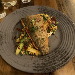 Trout - A dish with a seasoned fish fillet on a gray plate, accompanied by vegetables and grains, served in a restaurant setting.