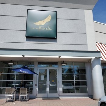 Mattarello - The photo depicts the exterior of Mattarello restaurant, featuring a modern facade with a sign displaying the restaurant's logo. The entrance has glass doors and large windows, with outdoor seating including a table, chairs, and a Peroni-branded umbrella. The building has a clean, contemporary design with neutral colors and is situated in a commercial area. - Italian Near Me - Markham, Ontario