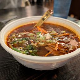 Sweet Potato Glass Noodles Soups - A bowl of hot and sour sweet potato noodle soup with beef, topped with chopped green onions, on a dark wooden table in a restaurant setting.