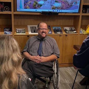 CoHatch - Me doing an interview in one of the meeting rooms within the offices of Sagamore Institute at CoHatch. - A man in a wheelchair, smiling, is engaged in conversation with two people in a room with bookshelves and a large screen. - Shared Office Spaces Near Me - Indianapolis, Indiana