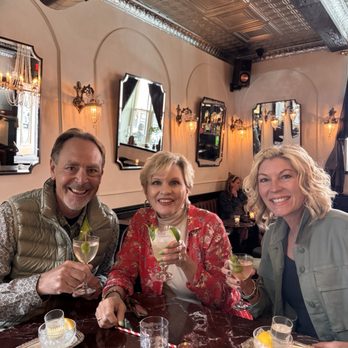 Van Atta - Three individuals sit at a marble-topped table in a vintage-inspired restaurant, holding cocktails with lime garnishes, smiling at the camera. - Bars Near Me - Telluride, Colorado