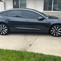 A black sedan with tinted windows is parked on a concrete driveway adjacent to a grassy area. The vehicle is positioned parallel to a residential building with large windows and a door, with a bicycle visible near the wall. The image captures the side profile of the car, showing its modern design and alloy wheels.