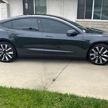 Rey's Window Tinting - A black sedan with tinted windows is parked on a concrete driveway adjacent to a grassy area. The vehicle is positioned parallel to a residential building with large windows and a door, with a bicycle visible near the wall. The image captures the side profile of the car, showing its modern design and alloy wheels. - Auto Glass Services Near Me - Brea, California