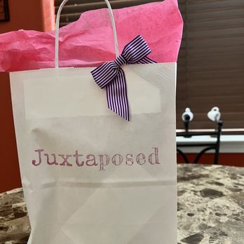 Juxtaposed - A white paper gift bag with the word 'Juxtaposed' printed in pink on the front, filled with pink tissue paper that extends above the bag's opening. A purple and white striped ribbon bow is attached to the upper part of the bag. The bag is placed on a marble table with a brown and beige pattern. In the background, there are window blinds and a pair of white earbuds resting on a black chair. - Home Decor Near Me - Albuquerque, New Mexico