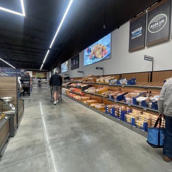 ALDI - Inside - The image shows the interior of an ALDI grocery store with a bakery section along the right side. Shelves are stocked with baked goods, and two customers are shopping, one pushing a cart and another carrying a blue shopping bag. The ceiling has modern lighting fixtures, and digital screens display food images and advertisements. The store layout is spacious with a polished concrete floor and clear signage. - Grocery Near Me - Wilmington, Delaware