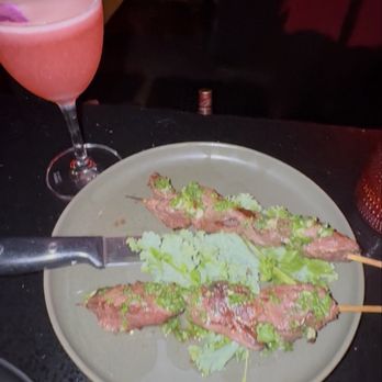 CODE RED - The image shows a dark table with a beige plate holding three skewers of grilled meat topped with green lettuce. A knife rests on the plate. To the left, there is a pink cocktail in a stemmed glass. The background is dimly lit, with some indistinct objects and a person partially visible. - Cocktail Bars Near Me - Washington, DC, District of Columbia