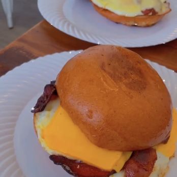 Scratch Kitchen - Single Hander sandwich with bacon - Two breakfast sandwiches are presented on white paper plates. Each sandwich contains a fried egg, melted cheese, and bacon, with toasted buns. The sandwiches are placed on a wooden table, with one sandwich in the foreground and the other slightly behind. The background shows part of a chair and a person’s hand near the second sandwich. - Breakfast & Brunch Near Me - Olney, Maryland