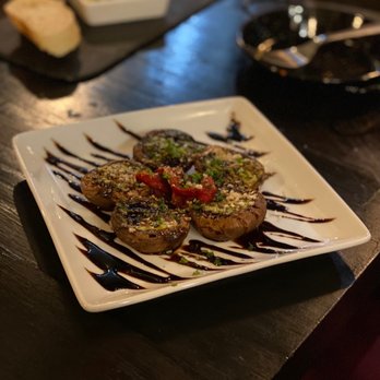 Debajo Tapas ABQ - Amazing mushroom dish! Champiñones al ajillo - A square white plate features stuffed mushrooms topped with chopped herbs and red pepper slices. The mushrooms are garnished with grated cheese or breadcrumbs and drizzled with balsamic glaze. The dish is presented on a dark wooden table, with a small bowl of olive oil and bread in the background, indicating a dining setting at a restaurant. - Spanish Near Me - Albuquerque, New Mexico