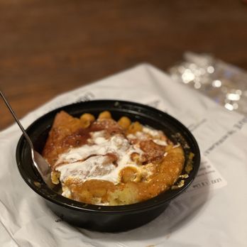 Indian Kitchen - Samosa Chaat - A black plastic bowl filled with samosa chaat, including pieces of samosa, chickpeas, yogurt, and sauce, is placed on a white paper on a wooden table. The background shows a blurred setting, likely inside a restaurant, with some reflective objects. The image captures a close-up of the dish, emphasizing the textures and toppings of the Indian snack. - Indian Near Me - Allston, Massachusetts