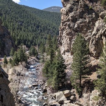Front Range Climbing Co - A natural mountain landscape shows a river flowing through a rocky canyon with large boulders along its banks. Tall pine trees grow on the canyon's sides, and dense forest covers the mountain slopes in the background. The sky is clear and blue, indicating a sunny day in a mountainous region. - Climbing Near Me - Colorado Springs, Colorado