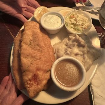 Skinflints - Fish and Chips - A plate features a large piece of battered fried fish, mashed potatoes, coleslaw, lemon wedges, and two dipping sauces, including tartar sauce and gravy. The meal is served on a white plate placed on a dark wooden table, with a person's hand visible holding the plate and another hand reaching for utensils. The setting appears to be a casual dining environment, possibly a pub or restaurant. - American Near Me - Brooklyn, New York