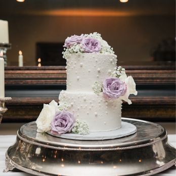 Villa de Amore - A two-tiered wedding cake decorated with purple and white roses, along with small white flowers, is displayed on a silver cake stand. The cake has a smooth white icing surface with small pearl-like decorations. The background includes a wooden surface and candles, indicating an indoor setting for a wedding celebration. - Venues & Event Spaces Near Me - Temecula, California
