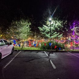 This is as you pull up . It's open until ten..So beautiful . - Outdoor area at night decorated with string lights, trees, and festive banners, with a parking lot in the foreground.