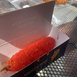 A mochi donut on a stick, coated with bright red crumbs, is positioned inside a black and white branded box on a metal outdoor table. In the background, there is a tray of fried food on a skewer and a clear plastic container, suggesting a food stall or outdoor dining setting. The image captures the presentation of a mochi donut at Mochi Ring, a dessert shop specializing in Asian fusion snacks.
