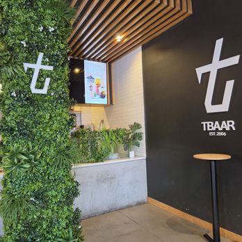Tbaar - Bayside - front counter - The photo depicts the interior of Tbaar, a business established in 2006. It includes a green wall with various plants and a white logo, a black wall with the Tbaar logo and establishment year, a wooden ceiling with slats, and a small round table. A digital menu display is visible near the counter area, which has a concrete finish and some potted plants. - Bubble Tea Near Me - Bayside, New York
