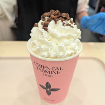 Molly Tea - Beautiful Jasmine milk tea with whip cream - A pink paper cup labeled 'Oriental Jasmine' holds jasmine milk tea topped with a generous swirl of whipped cream and sprinkled with chocolate shavings. The cup features a black floral logo and is placed on a light-colored surface. In the background, a person wearing a black apron and blue gloves is visible, indicating a cafe or tea shop setting. - Bubble Tea Near Me - Markham, Ontario
